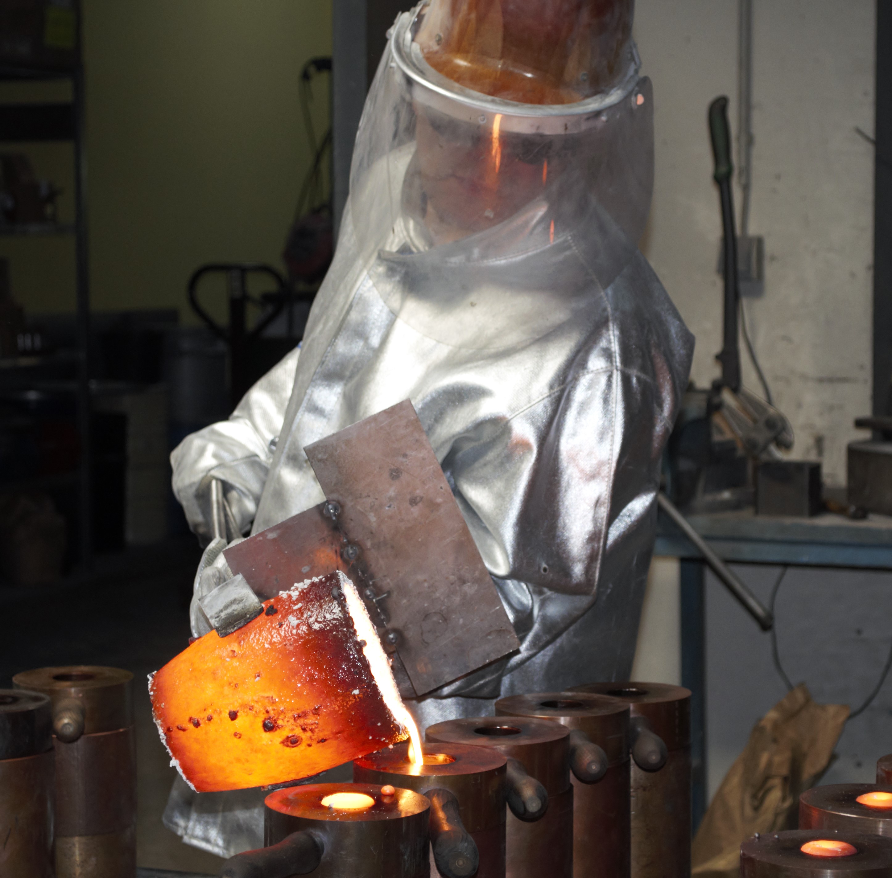 Zu sehen ist ein Mitarbeiter, gekleidet in vollständiger Schutzkleidung, wie er geschmolzenes Metall in die Formen gießt. 