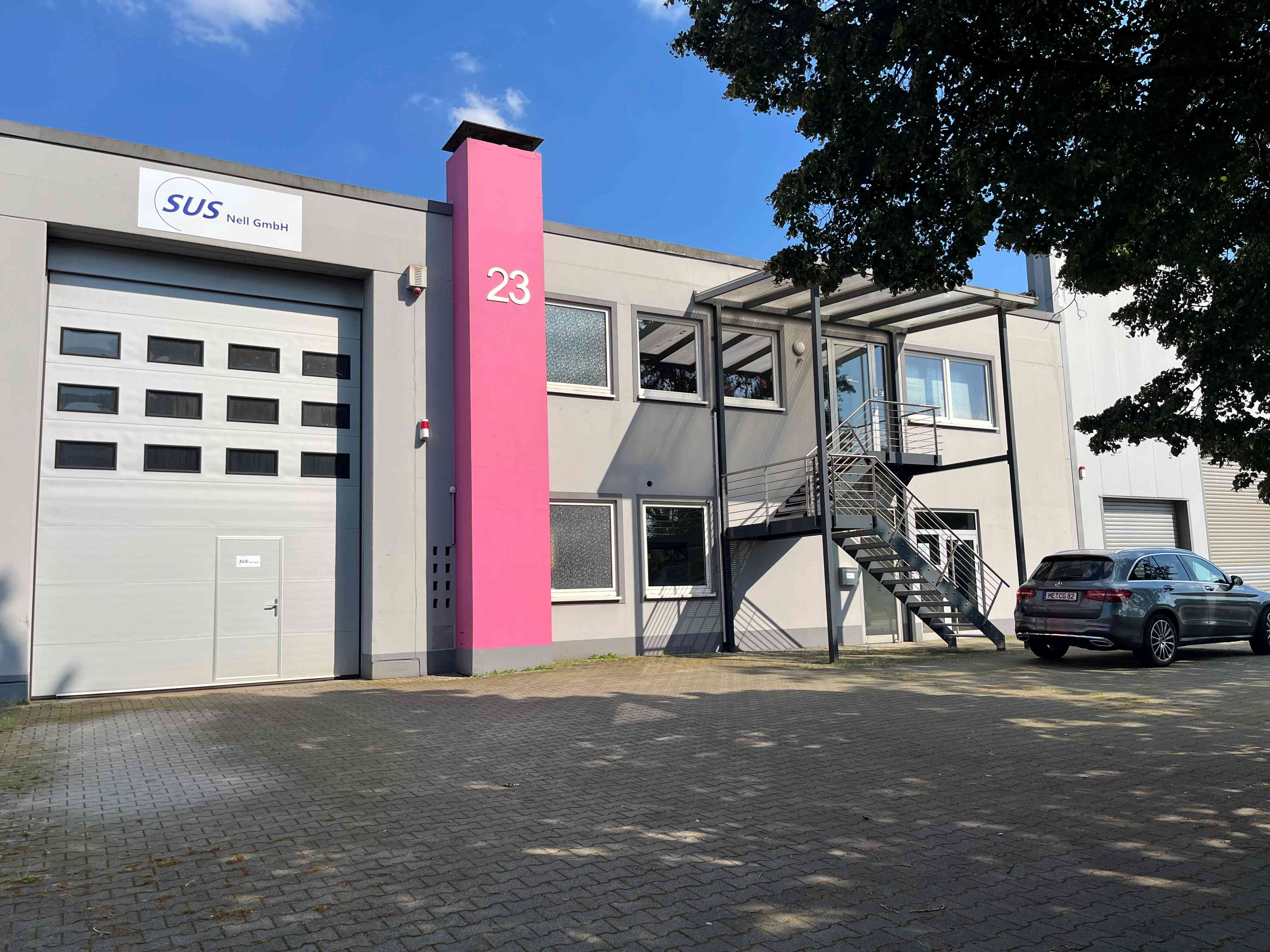 Zu sehen ist das Firmengebäude der SUS Nell GmbH. Ein großer roter Kamin mit einer 23 darauf steht in der mitte.